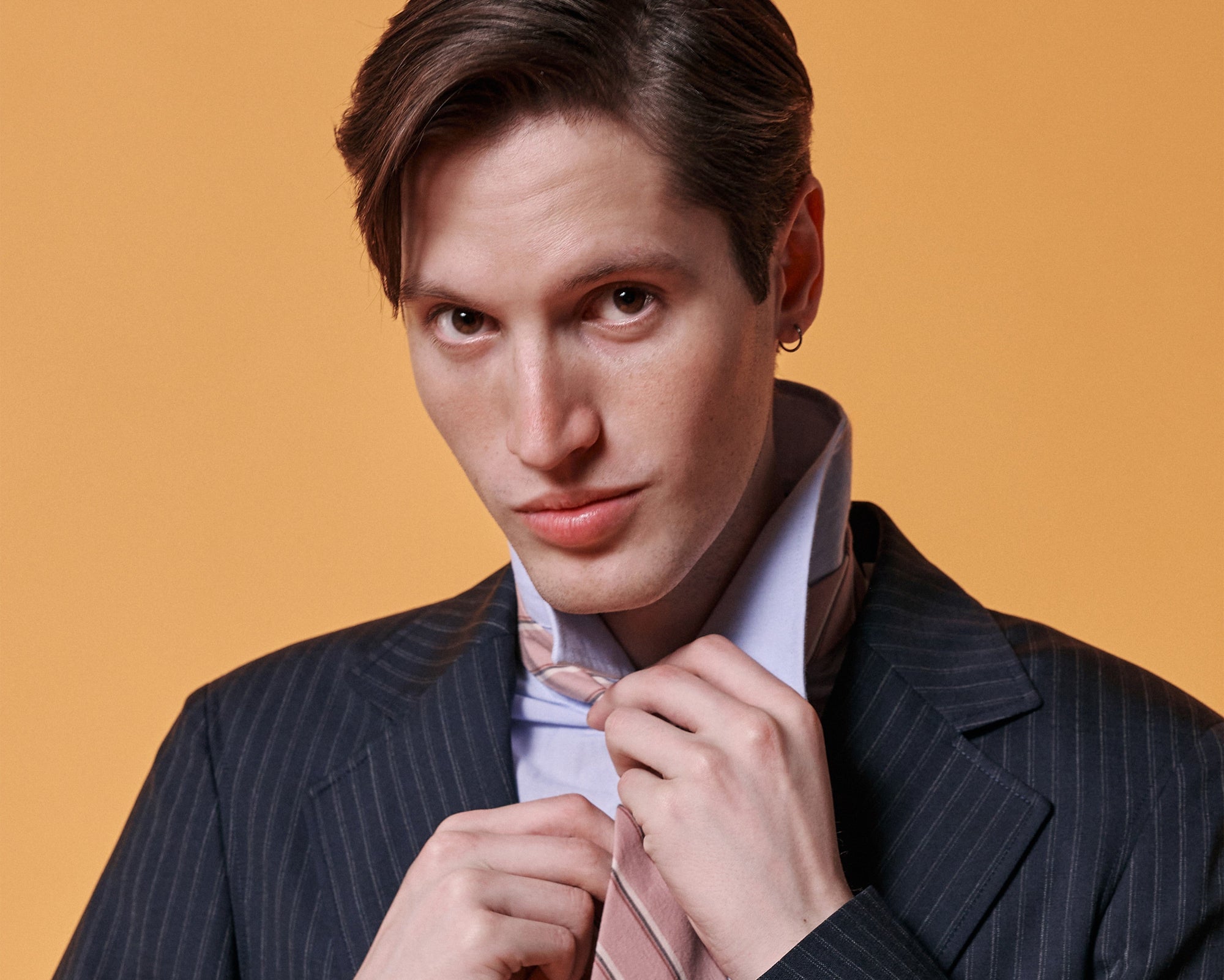 alt="Close-up of model wearing a navy pinstripe unstructured suit with a pale blue shirt and pink tie against a warm orange background
