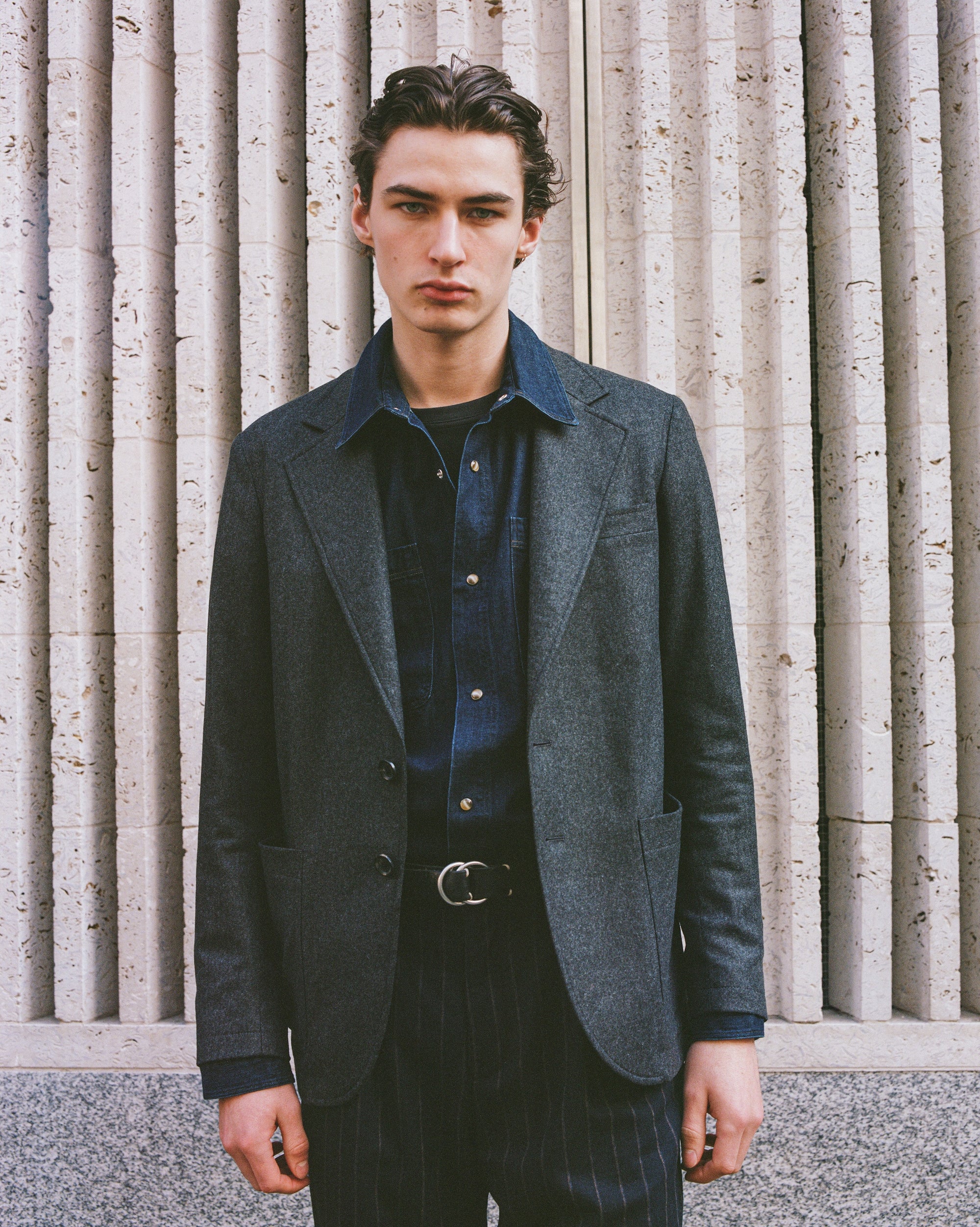 Model wearing a dark gray Oliver Spencer blazer over a blue shirt against a textured wall.
