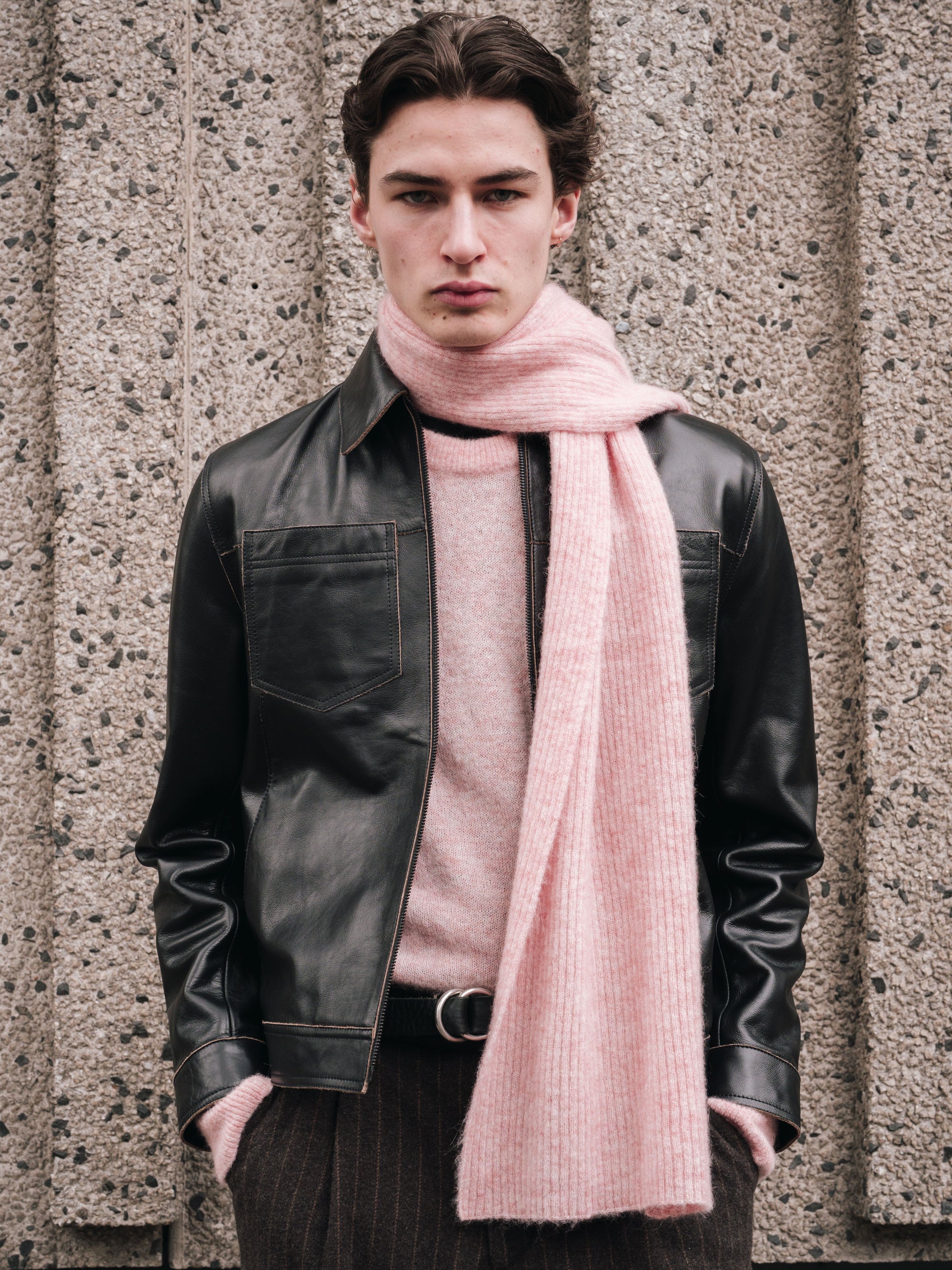 Model wearing a black Oliver Spencer leather jacket and pink scarf against a textured wall.