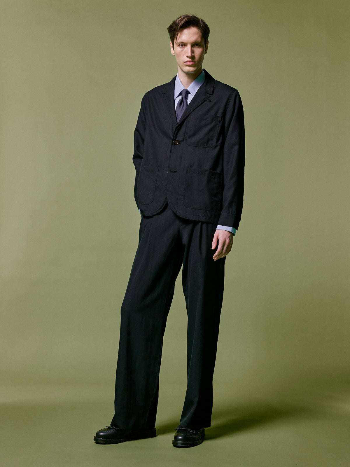 Black suit with a blue shirt and spotted tie, against an olive backdrop.