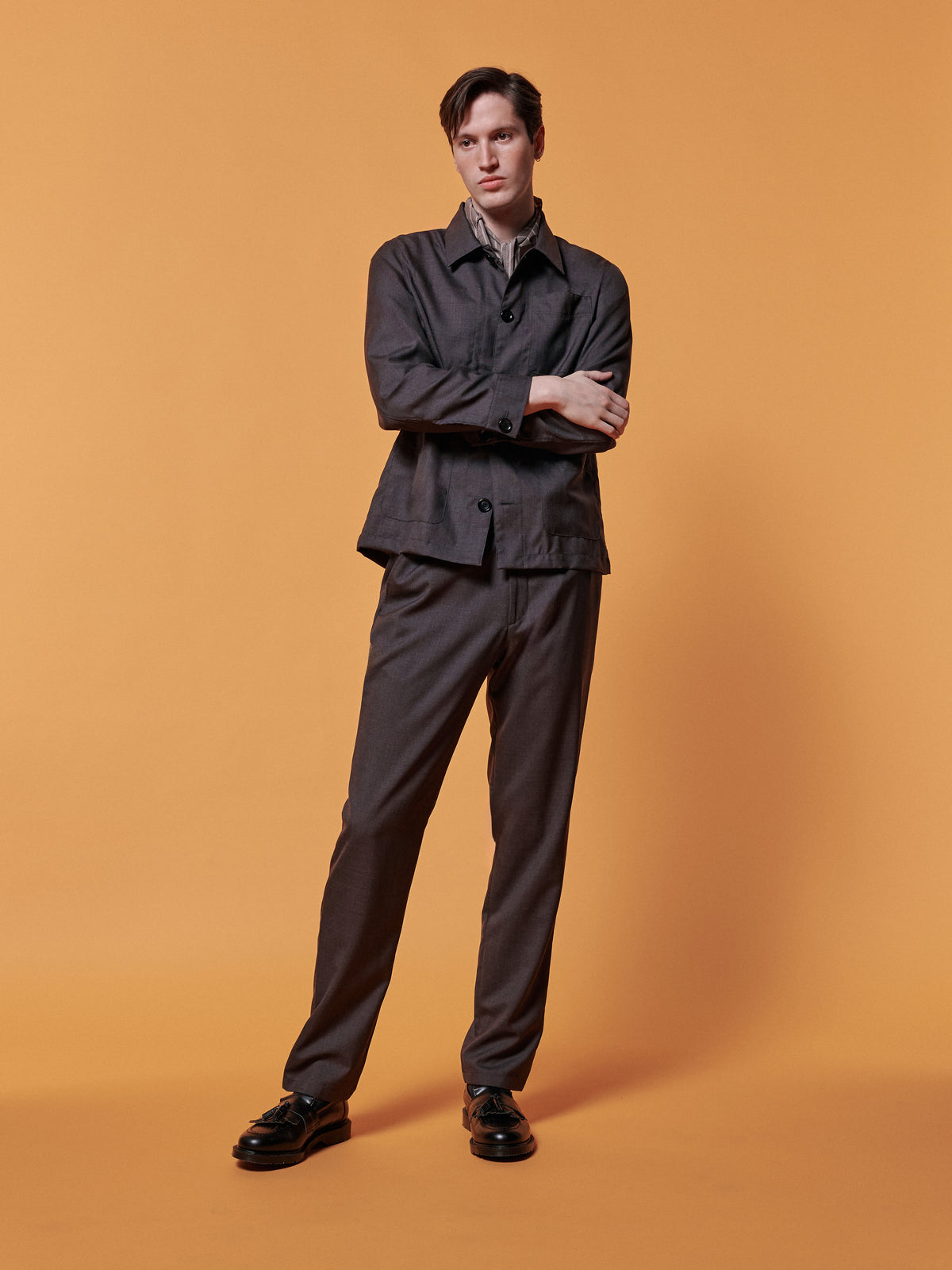 Brown jacket and trousers with a neckerchief, arms folded, against an ochre backdrop.