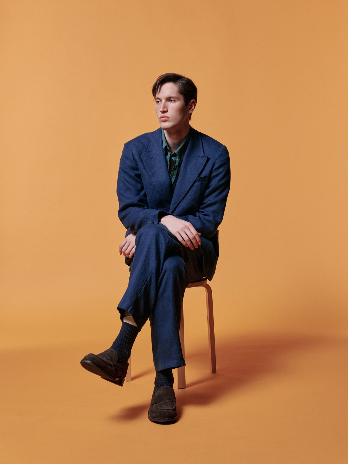 Navy suit, seated on a stool against an ochre backdrop.