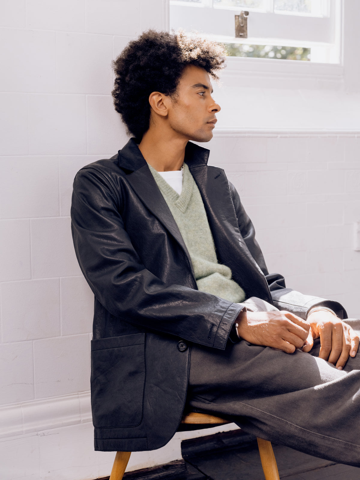 Black leather Westbury jacket over a pale green jumper with grey trousers, seated.