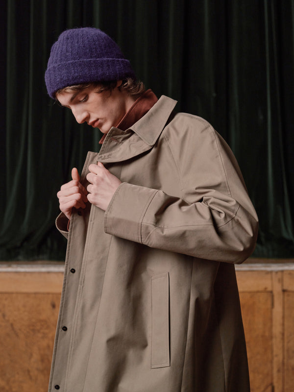 Man in a purple wool beanie fastening a stone cotton trench coat