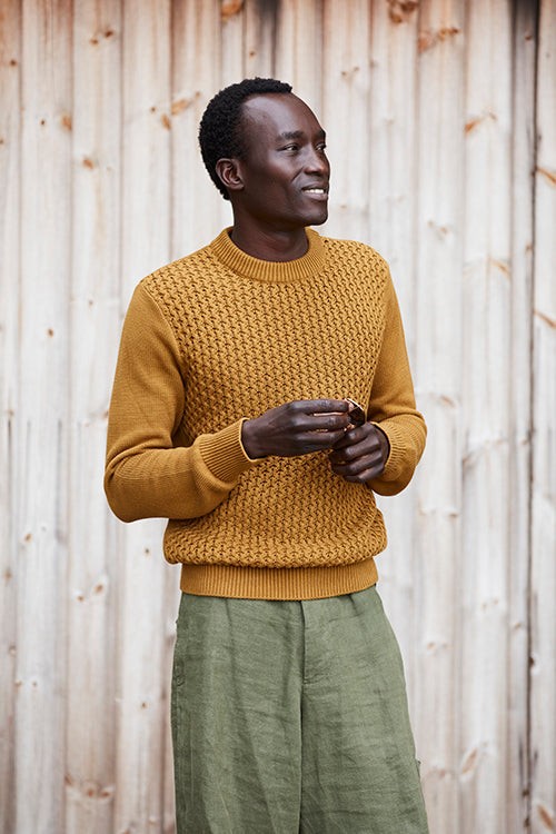Oliver Spencer Spring 21, look 2. Ochre ribbed jumper and green trousers against a slatted wooden wall.