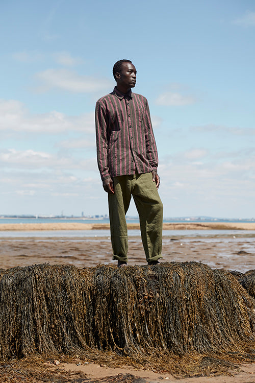 Oliver Spencer Spring 21, look 8. Rust check shirt and olive trousers, standing on seaweed-covered rock.