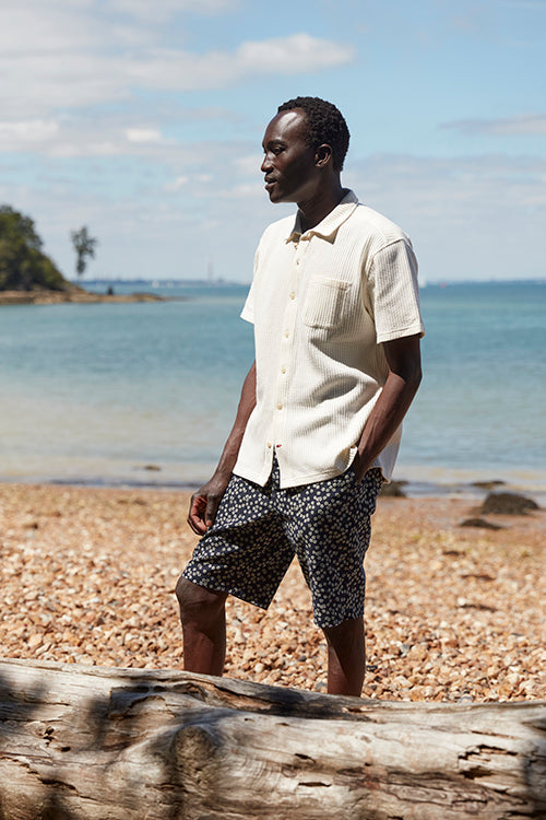 Oliver Spencer Spring 21, look 9. Cream short sleeve shirt and shorts, walking on driftwood.