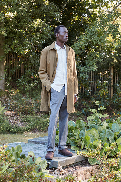 Oliver Spencer Spring 21, look 16. Camel overcoat over a pale shirt in a garden.