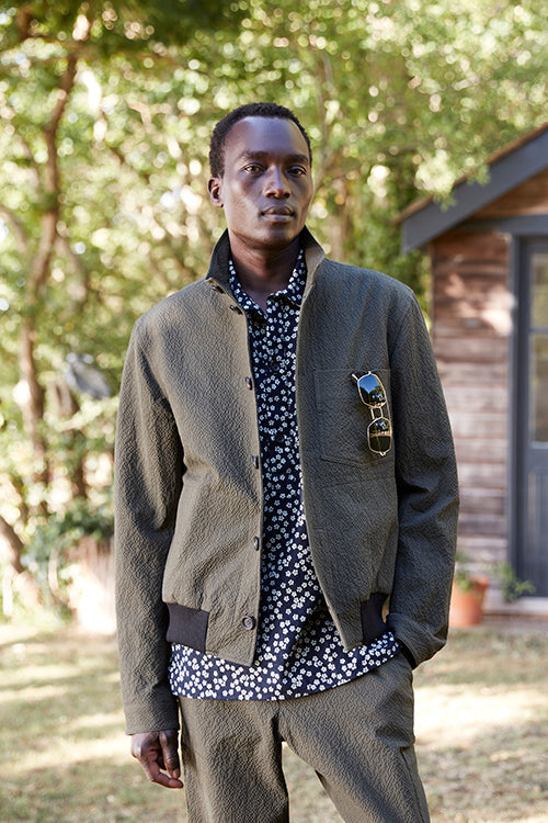 Oliver Spencer Spring 21, look 20. Olive jacket over a patterned navy shirt beside a wooden building.