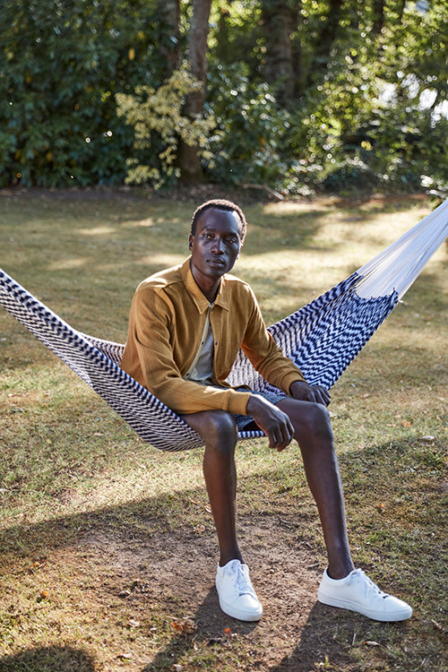 Oliver Spencer Spring 21, look 22. Seated in a hammock in an ochre jacket and grey trousers.