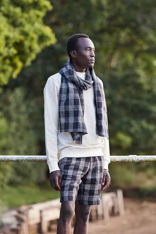 Oliver Spencer Spring 21, look 26. Cream jumper, navy scarf and navy check shorts beside water.