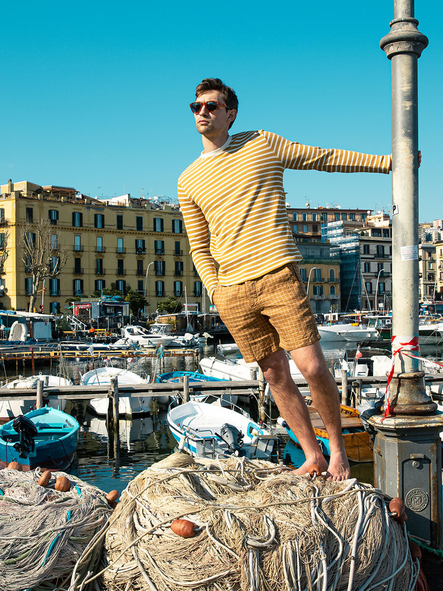 Oliver Spencer Summer 22 in Naples, look 2. Yellow striped knit and shorts on the harbour wall, boats behind.