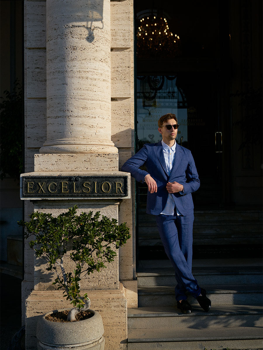 Oliver Spencer Summer 22 in Naples, look 4. Navy suit beside a stone column and a shop sign.