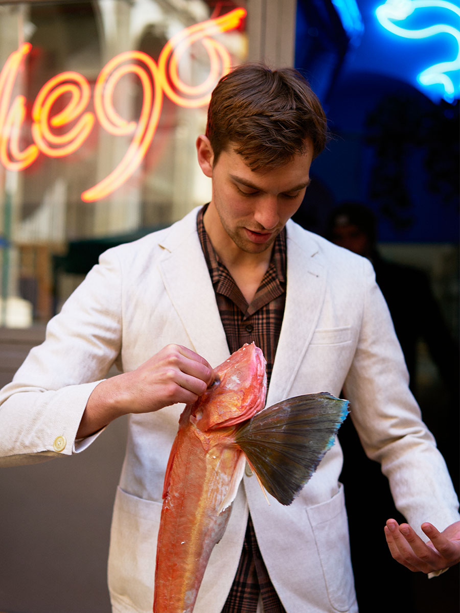 Oliver Spencer Summer 22 in Naples, look 5. White jacket and scarf under neon signs at night.