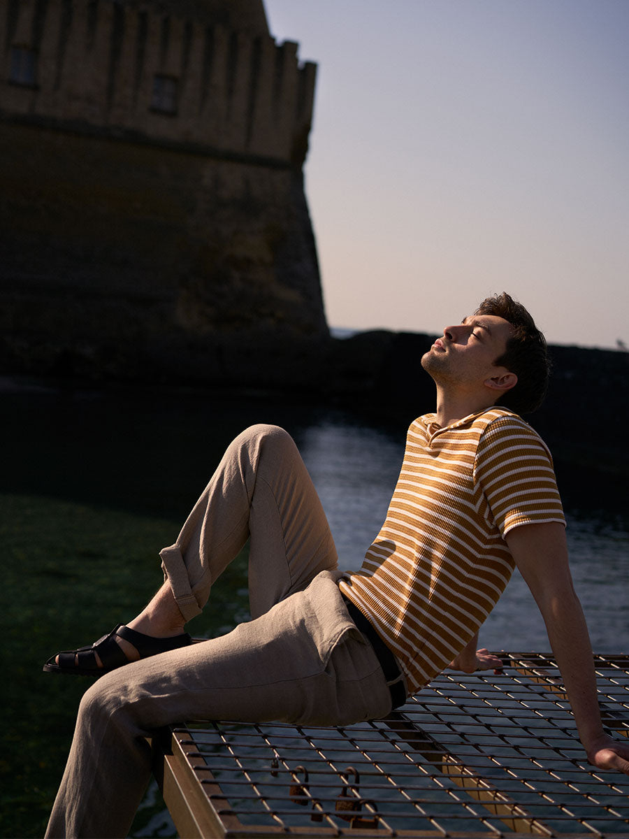Oliver Spencer Summer 22 in Naples, look 6. Striped knit, seated on stone steps at dusk with the castle behind.