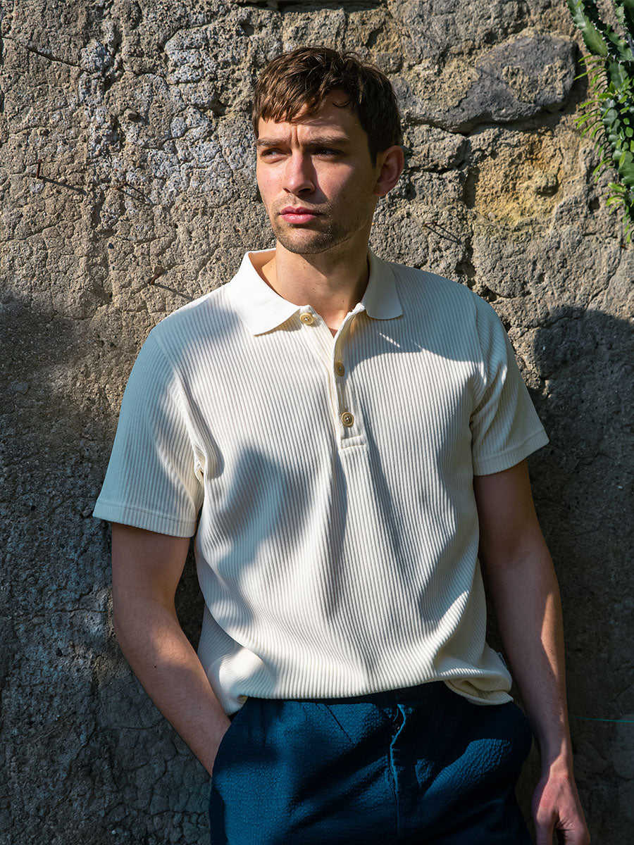 Oliver Spencer Summer 22 in Naples, look 11. Pale blue T-shirt and navy trousers against a stone wall.
