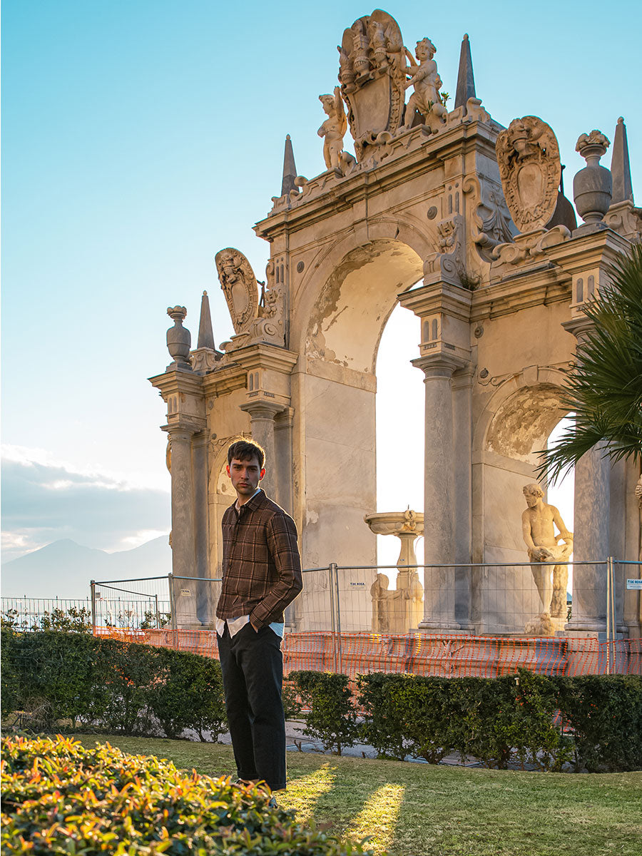 Oliver Spencer Summer 22 in Naples, look 12. Standing under a baroque arch with greenery behind.