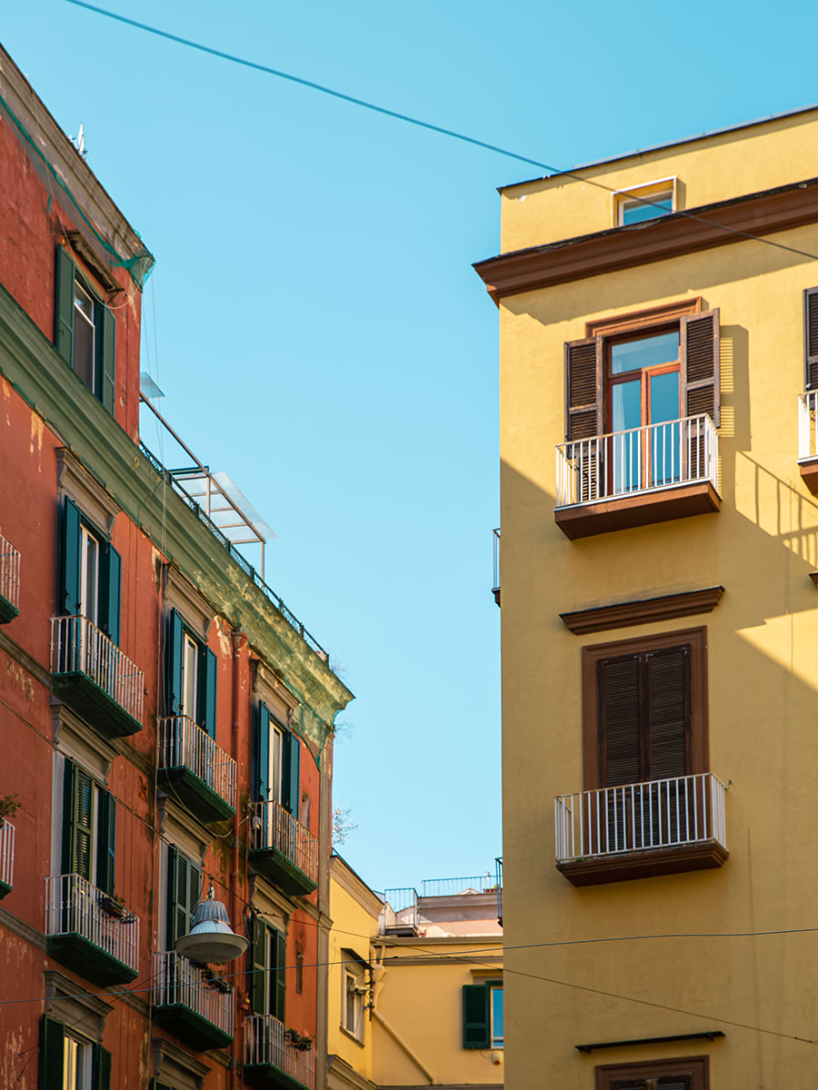 Oliver Spencer Summer 22 in Naples, look 17. Check jacket and trousers against a yellow building with balconies.