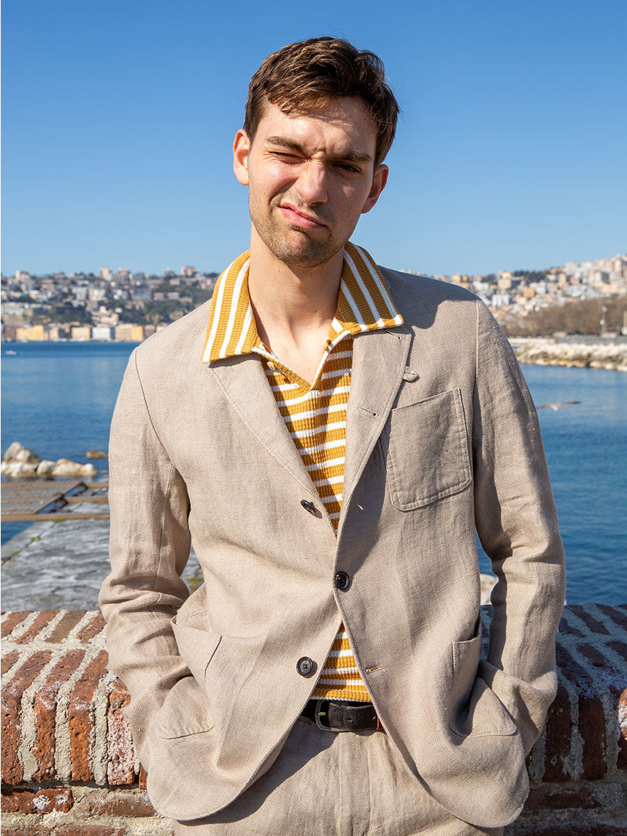 Oliver Spencer Summer 22 in Naples, look 18. Cream jacket over a yellow knit, the bay behind.