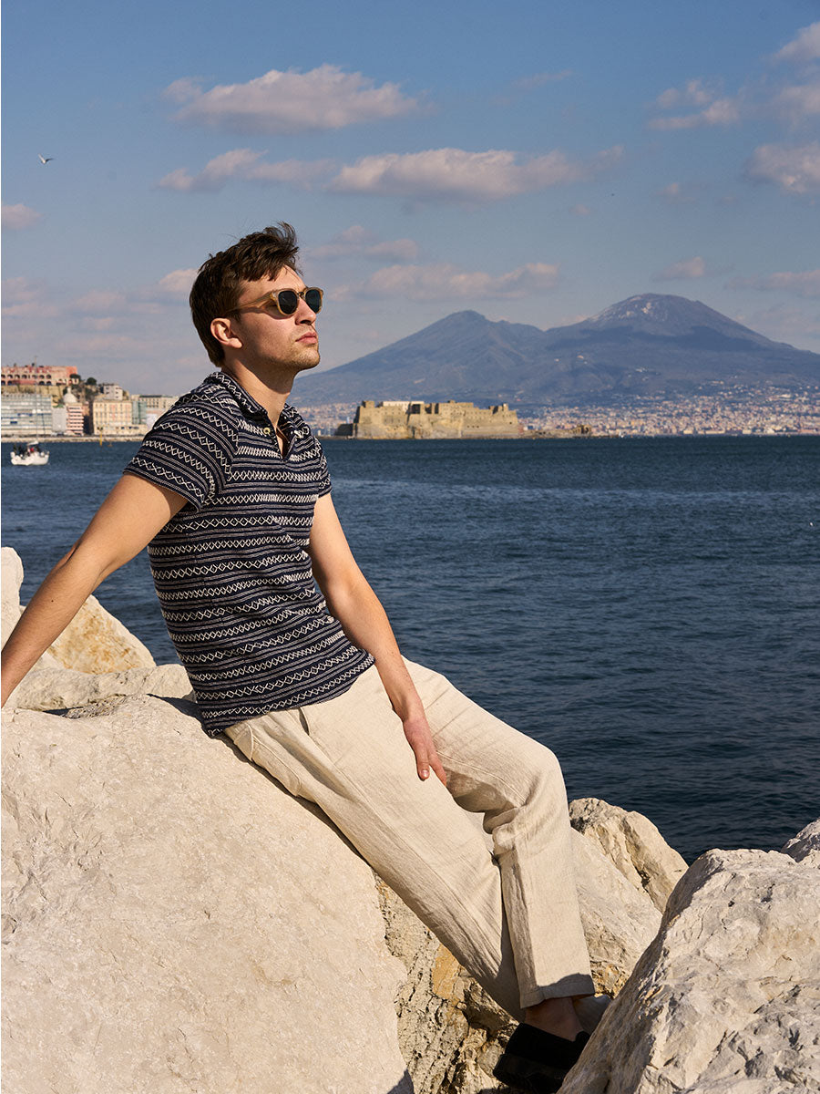 Oliver Spencer Summer 22 in Naples, look 19. Navy polo, seated on rocks with Vesuvius across the water.