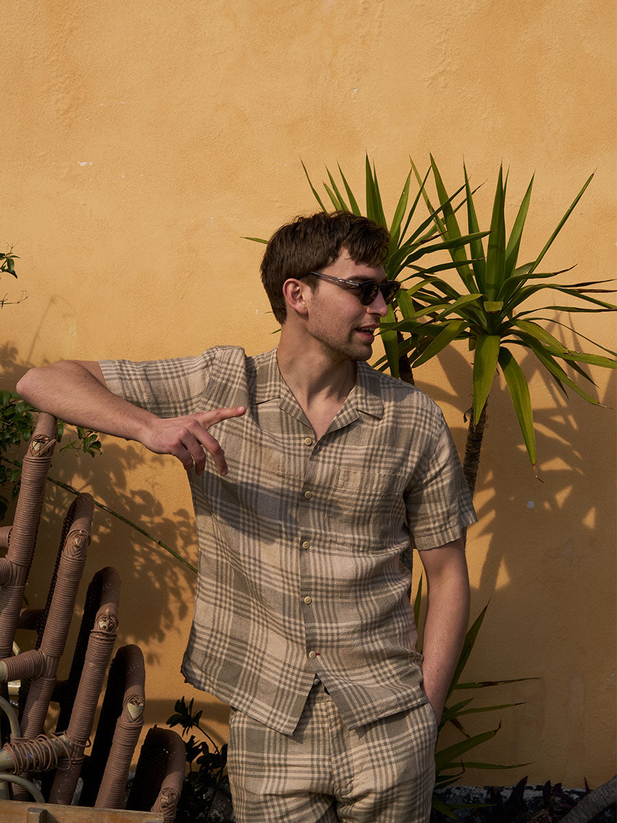 Oliver Spencer Summer 22 in Naples, look 24. Check shirt, seated among stacked cafe chairs and plants.